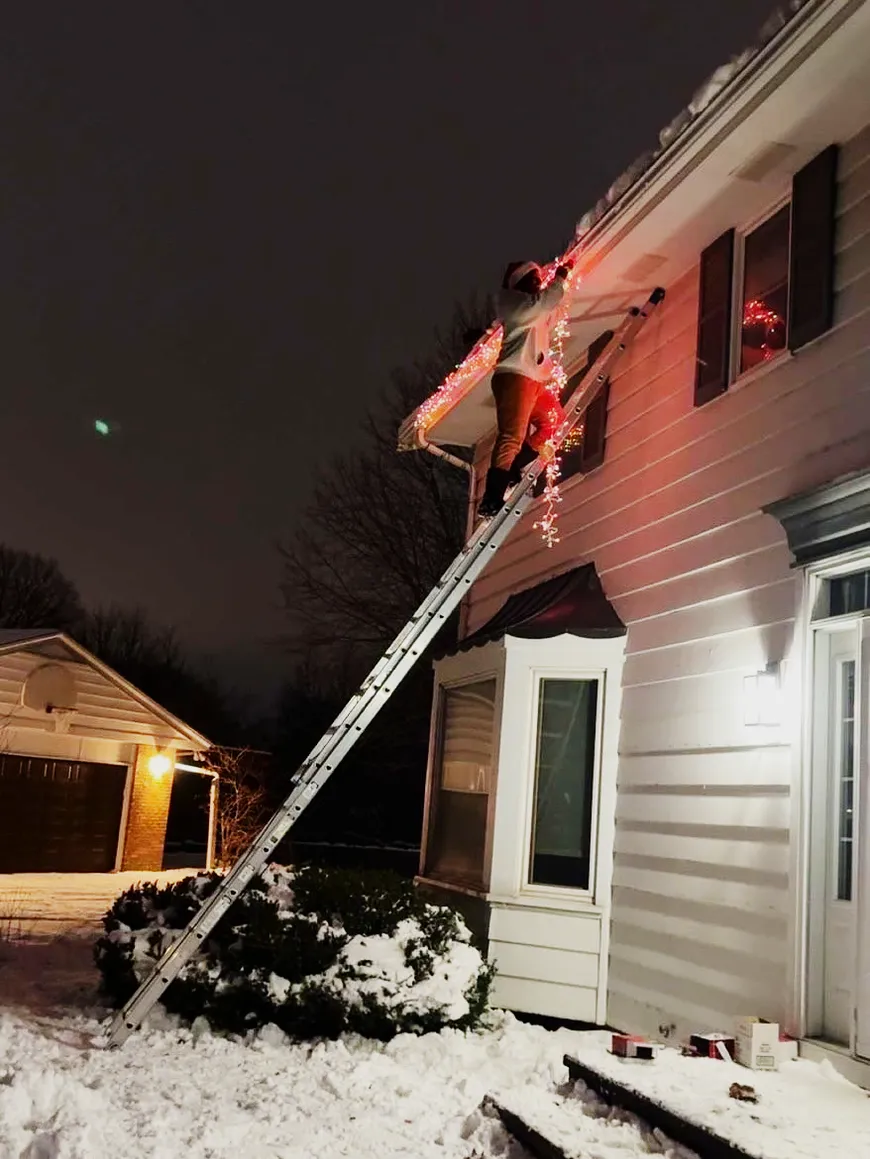 Maridadi installer on a tall ladder hanging holiday lights on a snowy evening